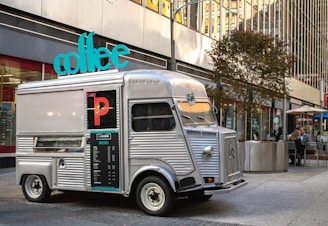 A vintage silver food truck with a large turquoise sign reading 'coffee' is parked on a city street. The truck features a menu on its side and several buildings reflecting in its windows. There are tables with people seated in the background, along with modern city architecture.