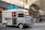 A vintage silver food truck with a large turquoise sign reading 'coffee' is parked on a city street. The truck features a menu on its side and several buildings reflecting in its windows. There are tables with people seated in the background, along with modern city architecture.