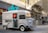 A vintage silver food truck with a large turquoise sign reading 'coffee' is parked on a city street. The truck features a menu on its side and several buildings reflecting in its windows. There are tables with people seated in the background, along with modern city architecture.