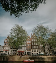 A picturesque row of traditional Dutch buildings line a canal. Lush green trees partially obscure the buildings, adding natural elements. Bicycles are parked along the canal, and a small red and white boat floats in the water. People are seen sitting by the water, creating a serene and lively atmosphere.