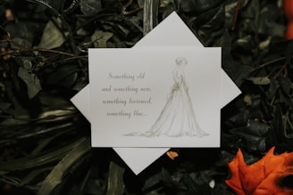A white card with elegant typography sits amidst dense green foliage. The card features the phrase 'Something old, something new, something borrowed, something blue...' accompanied by a light sketch of a bride in a flowing gown. There's an orange leaf to the side, adding a hint of color contrast.