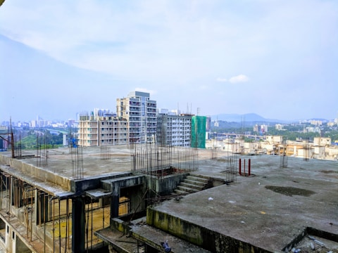 A partially constructed building with exposed steel rods, overlooking a cityscape with multiple high-rise buildings in the background. The construction site appears to be on a rooftop with a concrete surface, while green hills and an expansive sky provide a natural backdrop.