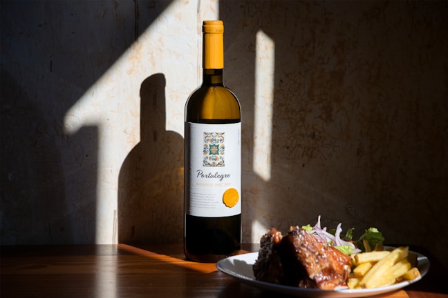 A bottle of wine is positioned upright on a wooden surface with a plate of food nearby, including ribs, fries, and a small salad. Sunlight creates dramatic shadows against a textured wall.