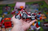 A hand holds a detailed toy motorcycle against a blurred background of colorful scattered toys on a play mat. The motorcycle is predominantly red with chrome accents, and it is centered in the image, drawing attention to its miniature size and intricate design.