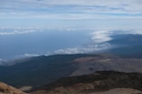 Aerial view of a vast landscape featuring a diverse terrain with mountains and forests. Above the terrain, a layer of clouds floats under a partly cloudy sky. The horizon shows a blend of sky and distant mountain ranges.