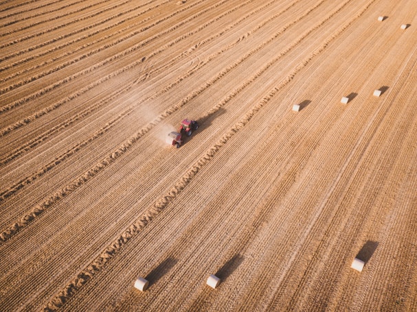 Aerial view of a large, flat agricultural field with a tractor harvesting crops. The field is divided into parallel rows with rectangular hay bales scattered across the landscape. Dust trails behind the moving tractor, highlighting the dry conditions.