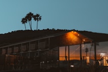 A building with large glass windows is illuminated by warm interior lighting, set against a backdrop of a hill and palm trees silhouetted in the evening sky. The structure appears to be a modern design with a mix of subtle stonework and smooth surfaces.