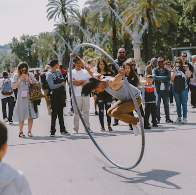 A performer in a gray tank top and brown pants is skillfully balancing inside a large ring, executing a stunt on a street. The action is closely observed by an audience gathered around, featuring people of various ages and expressions of curiosity and amazement. Palm trees are visible in the background, suggesting a sunny, outdoor setting.