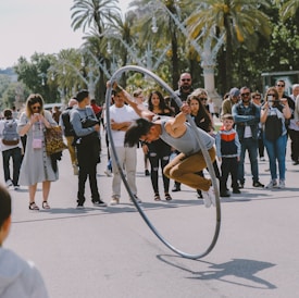 A performer in a gray tank top and brown pants is skillfully balancing inside a large ring, executing a stunt on a street. The action is closely observed by an audience gathered around, featuring people of various ages and expressions of curiosity and amazement. Palm trees are visible in the background, suggesting a sunny, outdoor setting.