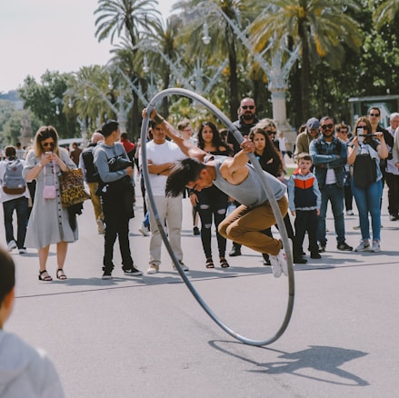 A performer in a gray tank top and brown pants is skillfully balancing inside a large ring, executing a stunt on a street. The action is closely observed by an audience gathered around, featuring people of various ages and expressions of curiosity and amazement. Palm trees are visible in the background, suggesting a sunny, outdoor setting.