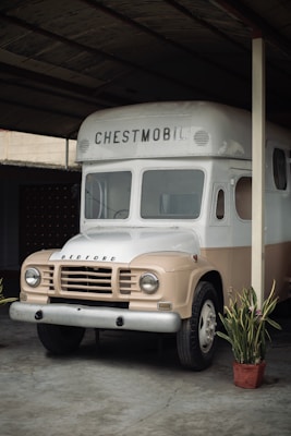 A vintage Bedford truck named 'Chestmobil' is parked under a shelter. The vehicle has a cream and beige color scheme with potted plants placed nearby, creating a nostalgic and historical atmosphere.