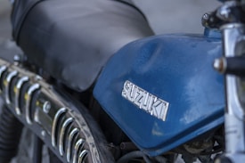 A close-up view of a blue Suzuki motorcycle tank with the brand logo prominently displayed. The image also shows parts of the seat and the side exhaust with chrome detailing.