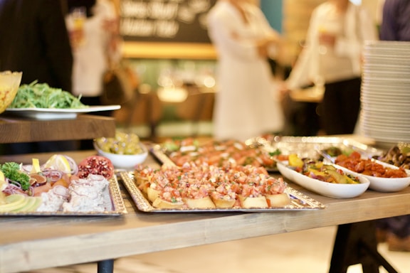 A beautifully arranged buffet table featuring various dishes including appetizers, salads, and small bites on trays and bowls. People are dressed in formal or semi-formal attire in the background, indicating a social gathering.