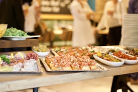 A beautifully arranged buffet table featuring various dishes including appetizers, salads, and small bites on trays and bowls. People are dressed in formal or semi-formal attire in the background, indicating a social gathering.