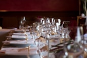 A beautifully set dining table with neatly arranged white napkins tied with beige ribbons. The table is adorned with sparkling glassware including wine glasses, suggesting a formal dining setting. Soft lighting enhances the elegant atmosphere.