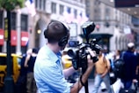 A person wearing headphones and a light blue shirt operates a professional video camera on a city street. American flags hang in the background above yellow taxis, suggesting an urban setting in the United States. Pedestrians and blurry motion add to the busy city atmosphere.