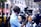 A person wearing headphones and a light blue shirt operates a professional video camera on a city street. American flags hang in the background above yellow taxis, suggesting an urban setting in the United States. Pedestrians and blurry motion add to the busy city atmosphere.