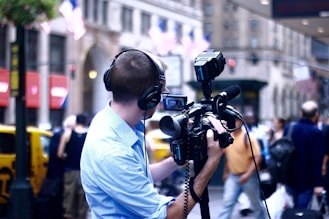 A person wearing headphones and a light blue shirt operates a professional video camera on a city street. American flags hang in the background above yellow taxis, suggesting an urban setting in the United States. Pedestrians and blurry motion add to the busy city atmosphere.
