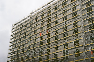 A large building under construction with extensive scaffolding covering its facade. The scaffolding includes metal poles and yellow safety barriers. Workers wearing bright orange clothing are visible on different levels, indicating active construction work.