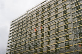 A large building under construction with extensive scaffolding covering its facade. The scaffolding includes metal poles and yellow safety barriers. Workers wearing bright orange clothing are visible on different levels, indicating active construction work.