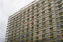 A large building under construction with extensive scaffolding covering its facade. The scaffolding includes metal poles and yellow safety barriers. Workers wearing bright orange clothing are visible on different levels, indicating active construction work.