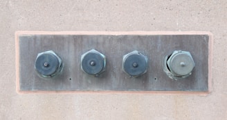 Four large metal bolts are embedded in a rectangular metal plate on a concrete wall. Each bolt has a round, flat head with a hexagonal base, showing signs of wear and tarnish.