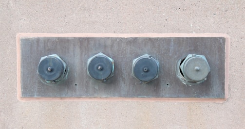 Four large metal bolts are embedded in a rectangular metal plate on a concrete wall. Each bolt has a round, flat head with a hexagonal base, showing signs of wear and tarnish.