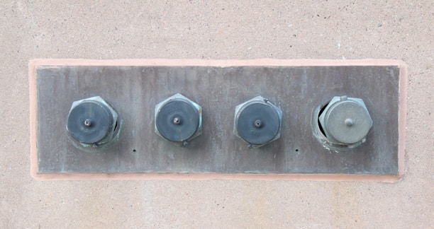 Four large metal bolts are embedded in a rectangular metal plate on a concrete wall. Each bolt has a round, flat head with a hexagonal base, showing signs of wear and tarnish.