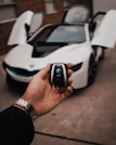 A hand holds a car key fob with a BMW logo prominently displayed. In the background, a white BMW sports car is parked with its gullwing doors open, creating an impression of luxury and modern design.