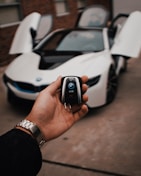 A hand holds a car key fob with a BMW logo prominently displayed. In the background, a white BMW sports car is parked with its gullwing doors open, creating an impression of luxury and modern design.