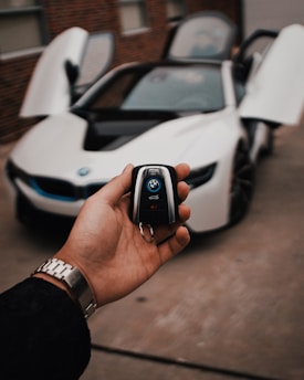A hand holds a car key fob with a BMW logo prominently displayed. In the background, a white BMW sports car is parked with its gullwing doors open, creating an impression of luxury and modern design.