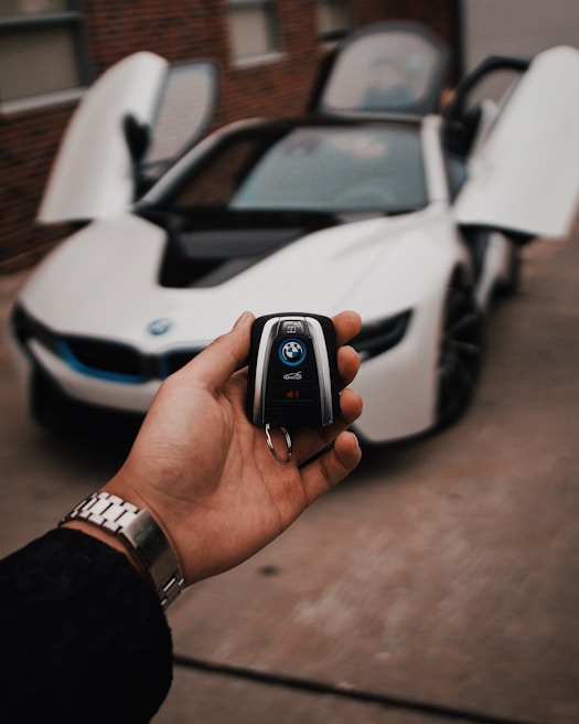A hand holds a car key fob with a BMW logo prominently displayed. In the background, a white BMW sports car is parked with its gullwing doors open, creating an impression of luxury and modern design.