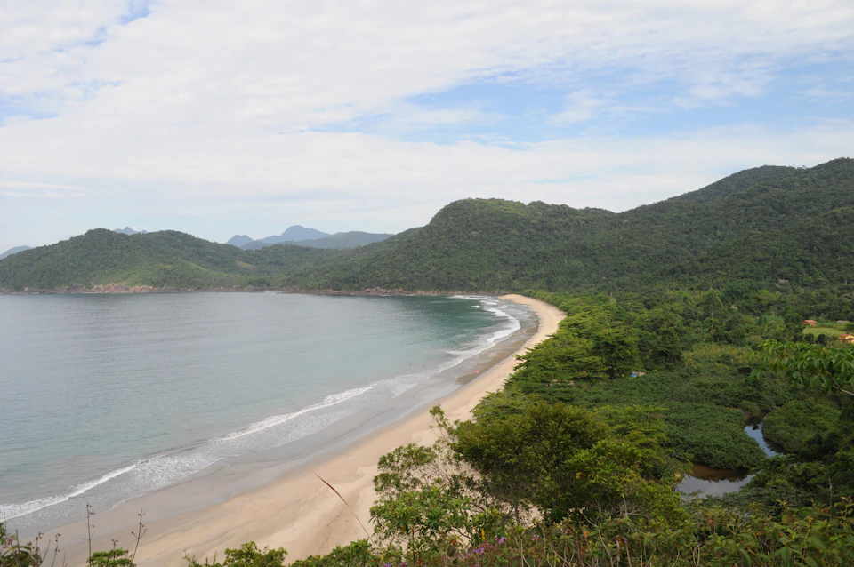 The image depicts a serene beach scene with a light beige sand beach stretching out towards the water, where small waves are gently lapping at the shoreline. The sky above is a clear blue, dotted with fluffy white clouds that add to the tranquil atmosphere of the scene. In the distance, there are mountains rising up against the horizon, their green and brown hues contrasting beautifully with the blue sky.