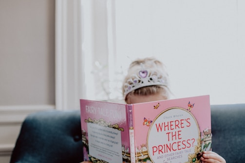 A child is sitting on a blue couch wearing a shiny tiara, absorbed in reading a pink book titled 'Where's the Princess?'.