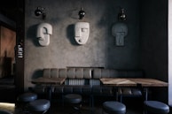 A modern interior space featuring a minimalist design with sculptural wall art. Three abstract, geometric sculptures resembling human faces are mounted on a textured gray wall. Below them is a leather bench sofa, accompanied by wooden tables and round, upholstered stools. The lighting is dim, creating an intimate ambiance.