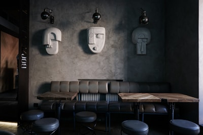 A modern interior space featuring a minimalist design with sculptural wall art. Three abstract, geometric sculptures resembling human faces are mounted on a textured gray wall. Below them is a leather bench sofa, accompanied by wooden tables and round, upholstered stools. The lighting is dim, creating an intimate ambiance.