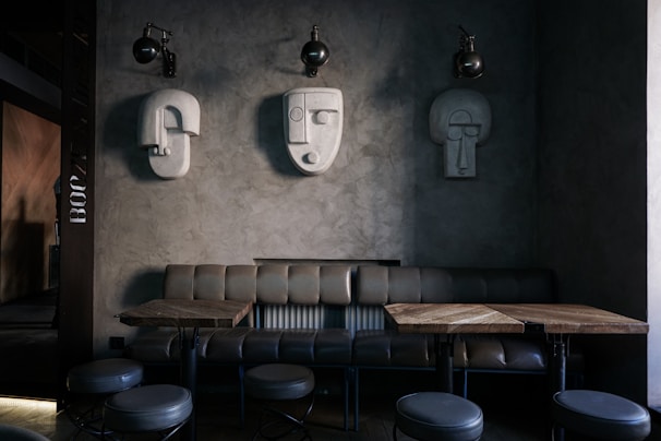 A modern interior space featuring a minimalist design with sculptural wall art. Three abstract, geometric sculptures resembling human faces are mounted on a textured gray wall. Below them is a leather bench sofa, accompanied by wooden tables and round, upholstered stools. The lighting is dim, creating an intimate ambiance.