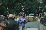 A person in a blue Lakers hoodie stands on a sidewalk in an urban park setting. The sidewalk is bustling with people, and in the background, there are trees and street signs. Another person is cycling on the path, and several people walk in different directions.