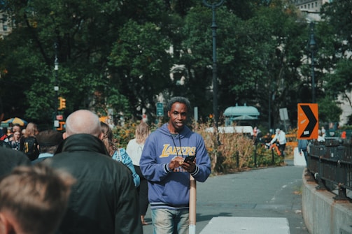 A person in a blue Lakers hoodie stands on a sidewalk in an urban park setting. The sidewalk is bustling with people, and in the background, there are trees and street signs. Another person is cycling on the path, and several people walk in different directions.