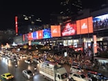 A busy urban street scene at night featuring heavy traffic with various vehicles including cars, taxis, and motorbikes. Pedestrians are gathered near a large building adorned with bright, colorful digital billboards and advertisements displaying dynamic visuals. The setting is lively and bustling.
