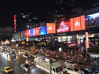 A busy urban street scene at night featuring heavy traffic with various vehicles including cars, taxis, and motorbikes. Pedestrians are gathered near a large building adorned with bright, colorful digital billboards and advertisements displaying dynamic visuals. The setting is lively and bustling.