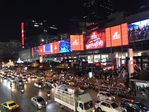A busy urban street scene at night featuring heavy traffic with various vehicles including cars, taxis, and motorbikes. Pedestrians are gathered near a large building adorned with bright, colorful digital billboards and advertisements displaying dynamic visuals. The setting is lively and bustling.