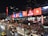 A busy urban street scene at night featuring heavy traffic with various vehicles including cars, taxis, and motorbikes. Pedestrians are gathered near a large building adorned with bright, colorful digital billboards and advertisements displaying dynamic visuals. The setting is lively and bustling.