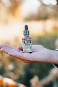 A person holds a small brown bottle labeled as 'Hemp Extract' with a dropper lid on an open palm. The background is softly blurred, suggesting an outdoor, nature setting.