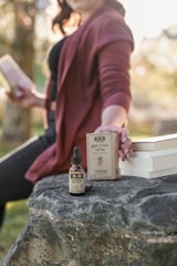 A person is sitting outdoors on a large rock, holding a box labeled 'Hemp Extract Tincture'. A bottle of tincture is placed on the rock. The person is wearing a maroon cardigan over a black top and holding a stack of books in the other hand. The setting appears to be in a natural, sunny environment with blurred tree foliage in the background.