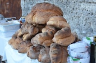A large stack of freshly baked, rustic bread loaves is arranged on a table. The loaves are irregularly shaped with a dark, crusty exterior, indicative of artisanal baking. Beside the bread, there are various kitchen items, including a bottle of olive oil, packs of paper plates, and plastic containers. The setup is against a rough stone wall, suggesting an outdoor or market setting.