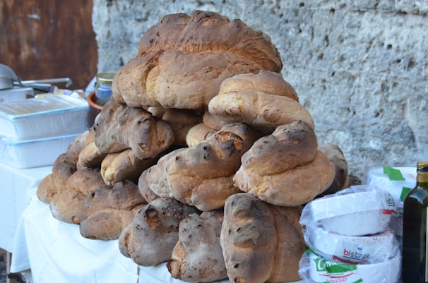 A large stack of freshly baked, rustic bread loaves is arranged on a table. The loaves are irregularly shaped with a dark, crusty exterior, indicative of artisanal baking. Beside the bread, there are various kitchen items, including a bottle of olive oil, packs of paper plates, and plastic containers. The setup is against a rough stone wall, suggesting an outdoor or market setting.