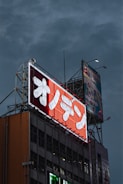 A large billboard with bright neon lights featuring Japanese characters is mounted on top of a building. The sign is primarily red and white, standing out against a cloudy, dark sky. Another billboard showing colorful graphics is also visible in the background.
