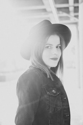 A monochrome portrait of a person wearing a wide-brimmed hat and a denim jacket. The lighting creates a soft, ethereal effect with noticeable lens flare and highlights, giving the scene a dreamy quality.
