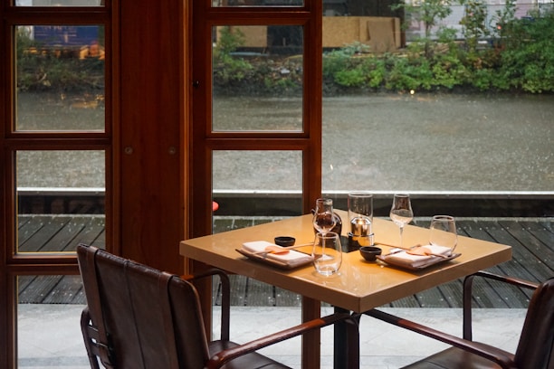 A cozy restaurant setting features a square table set for two, positioned next to large windows overlooking a body of water. The table is elegantly arranged with glassware, napkins, and small dishes, creating an inviting atmosphere. Outside, greenery and the reflection of the sky are visible, enhancing the serene ambiance.